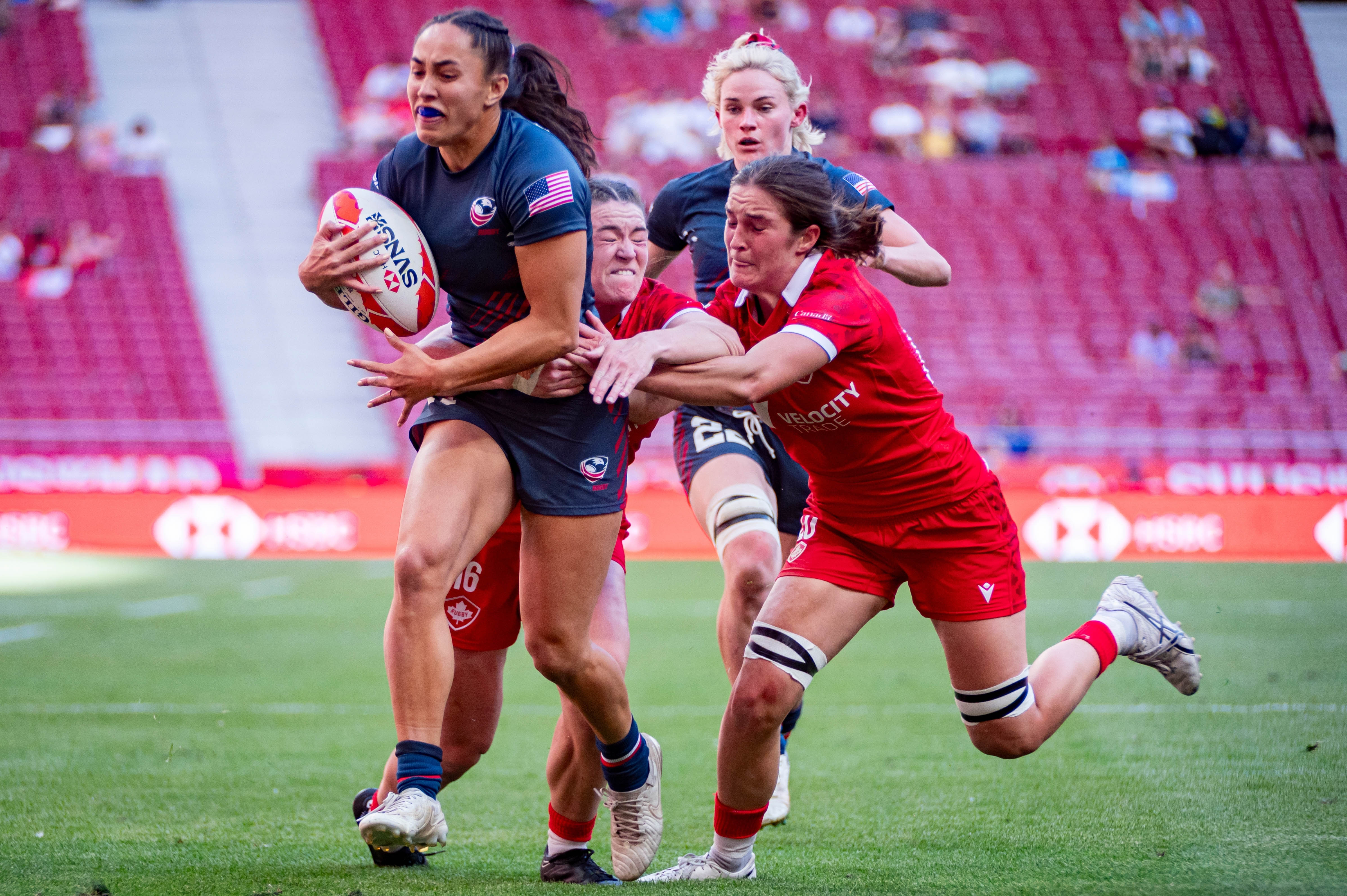 Rugby game at the Olympics - four women running, one with the ball, two attempting to grab her for a tackle, and a fourth chasing behind. Woman with the ball is an American athlete.